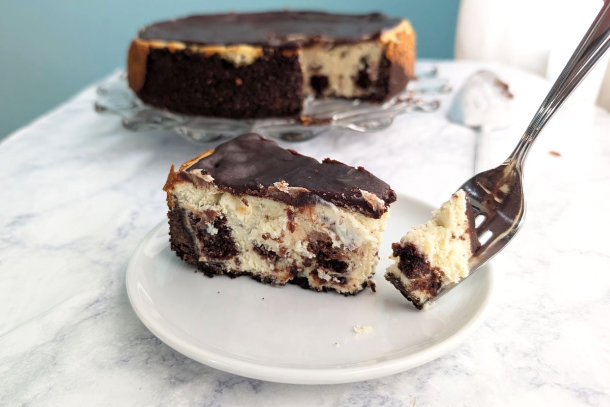 A bite of brownie cheesecake on a fork next to the slice of cheesecake. The rest of the cheesecake is in the background.