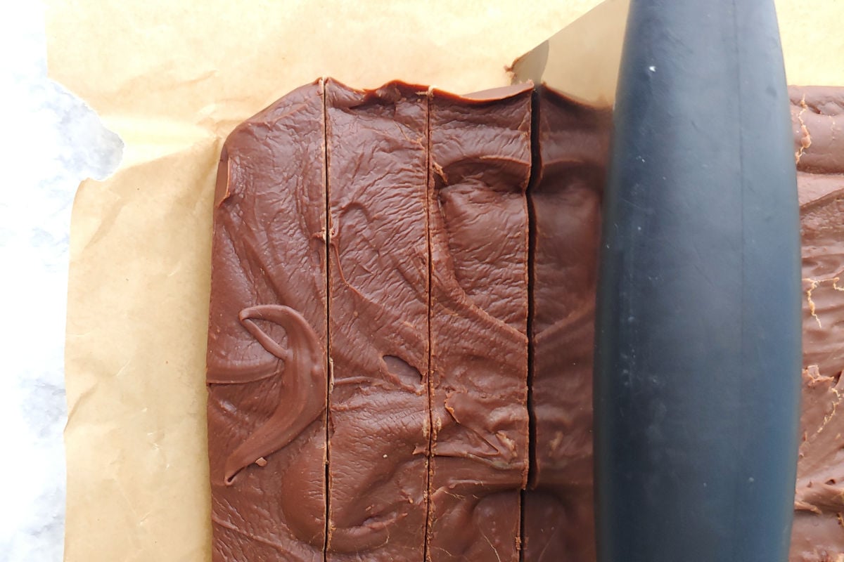 Chocolate fudge, on parchment paper on a counter, is cut into neat rows with a bench scraper.