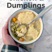 A hand places a bowl of slow cooker chicken and dumplings on top of a napkin. A bite of dumpling sits in the spoon in the bowl. Text overlay reads "Slow Cooker Chicken & Dumplings. Mostly Hands-Off. Easy As Cookies."