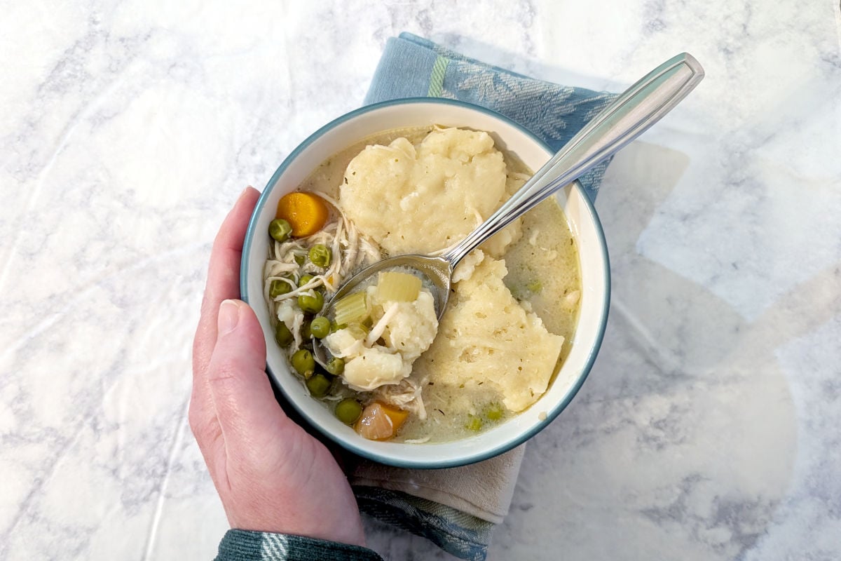 A hand places a bowl of slow cooker chicken and dumplings on top of a napkin. A bite of dumpling sits in the spoon in the bowl.