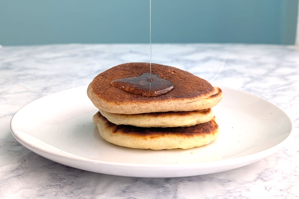 Syrup drizzles onto a stack of 3 pancakes on a plate.