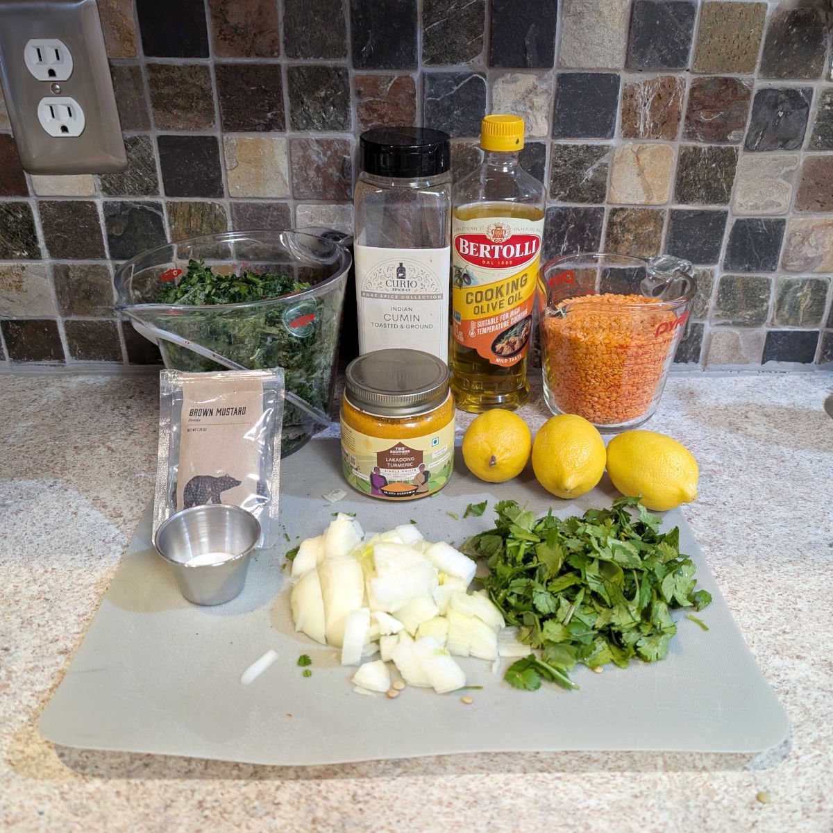 Ingredients for lemony lentil soup: a measuring cup of frozen chopped spinach, cumin, olive oil, a measuring cup of red lentils, mustard seeds, salt, turmeric, lemons, onion, and chopped cilantro.