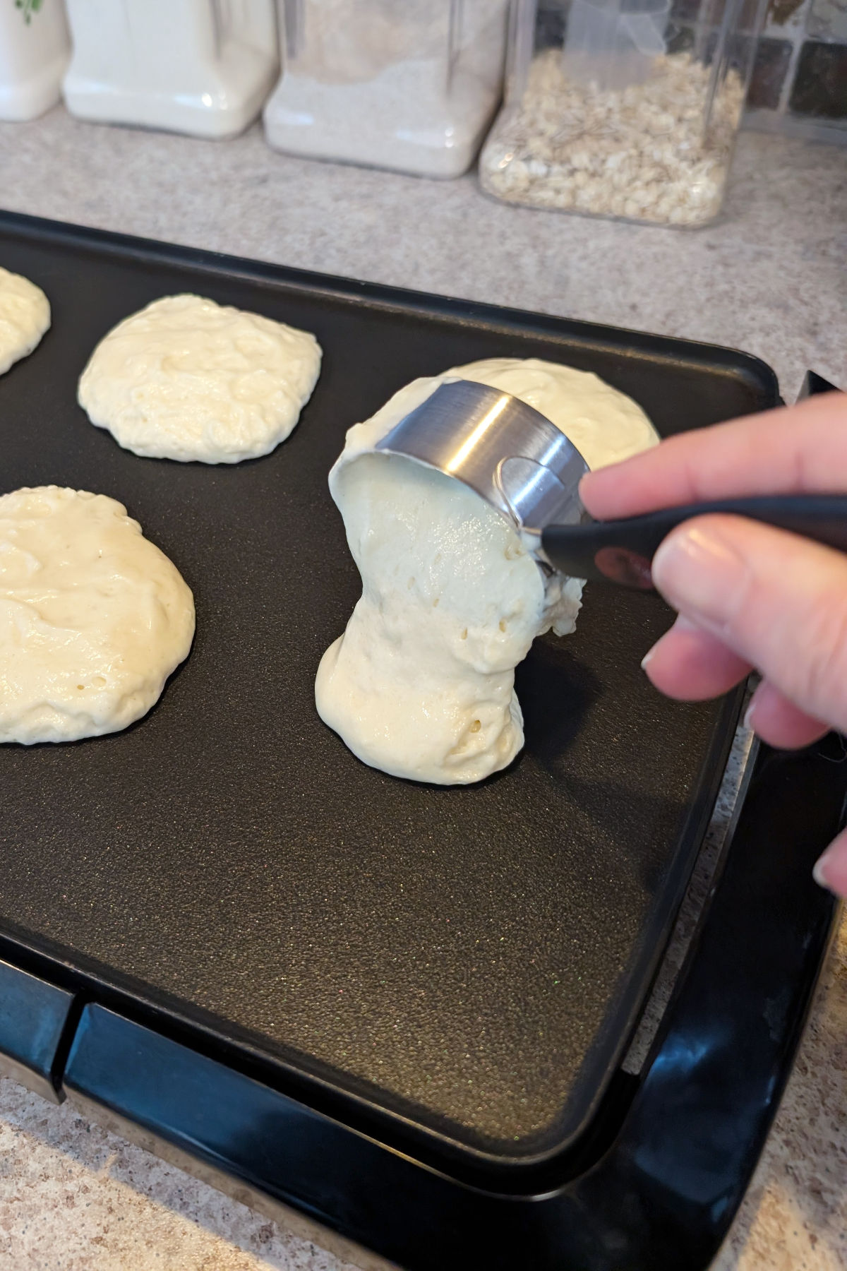 A hand pours pancake batter from a ¼ cup measuring cup onto a hot nonstick griddle. Other mounds of pancake batter are on the griddle.