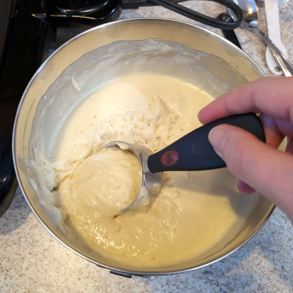 A hand digs a ¼ cup measuring scoop into a bowl of pancake batter, next to a nonstick griddle.