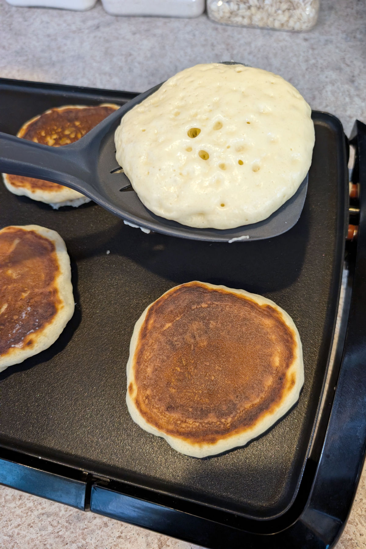 A flipping spatula holds a pancake over a griddle. The top of the pancake is still wet and uncooked. Flipped pancakes are on the nonstick griddle.