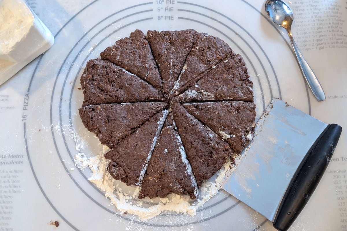 Double chocolate scone dough, cut into 10 wedges on a floured surface. A bench scraper rests next to the dough.