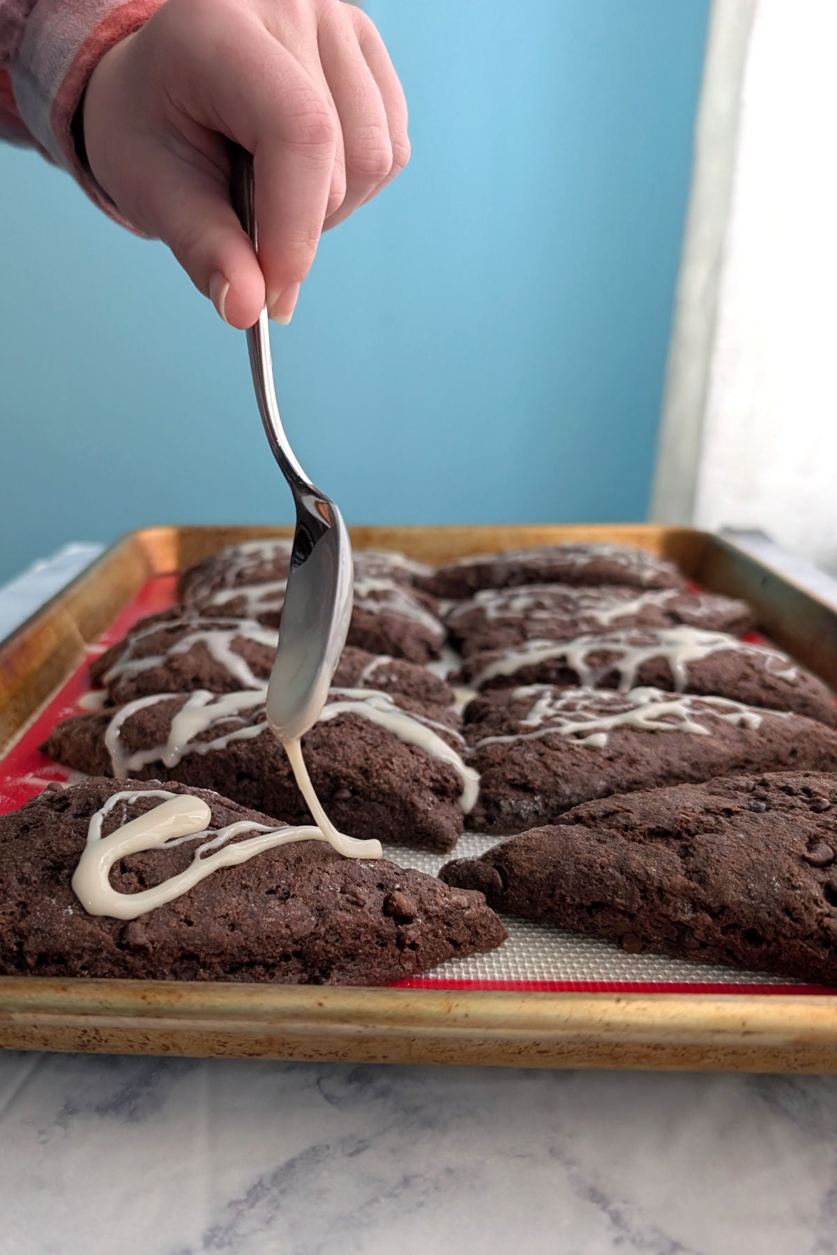 A hand holds a spoon drizzling white glaze onto double chocolate scones on a baking tray.