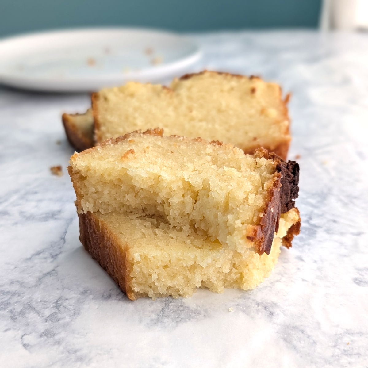 A slice of ricotta pound cake, broken into two and stacked to show texture. Background shows another slice of pound and a plate.