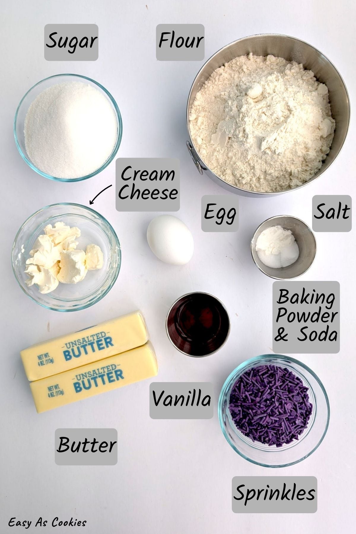 A flat-lay ingredient guide for sprinkle cookies on a white background. Labeled grey boxes identify bowls of flour, sugar, purple sprinkles, and cream cheese, along with two sticks of unsalted butter, an egg, vanilla extract, salt, and baking powder. A small arrow points from the cream cheese label to its bowl for clarity.