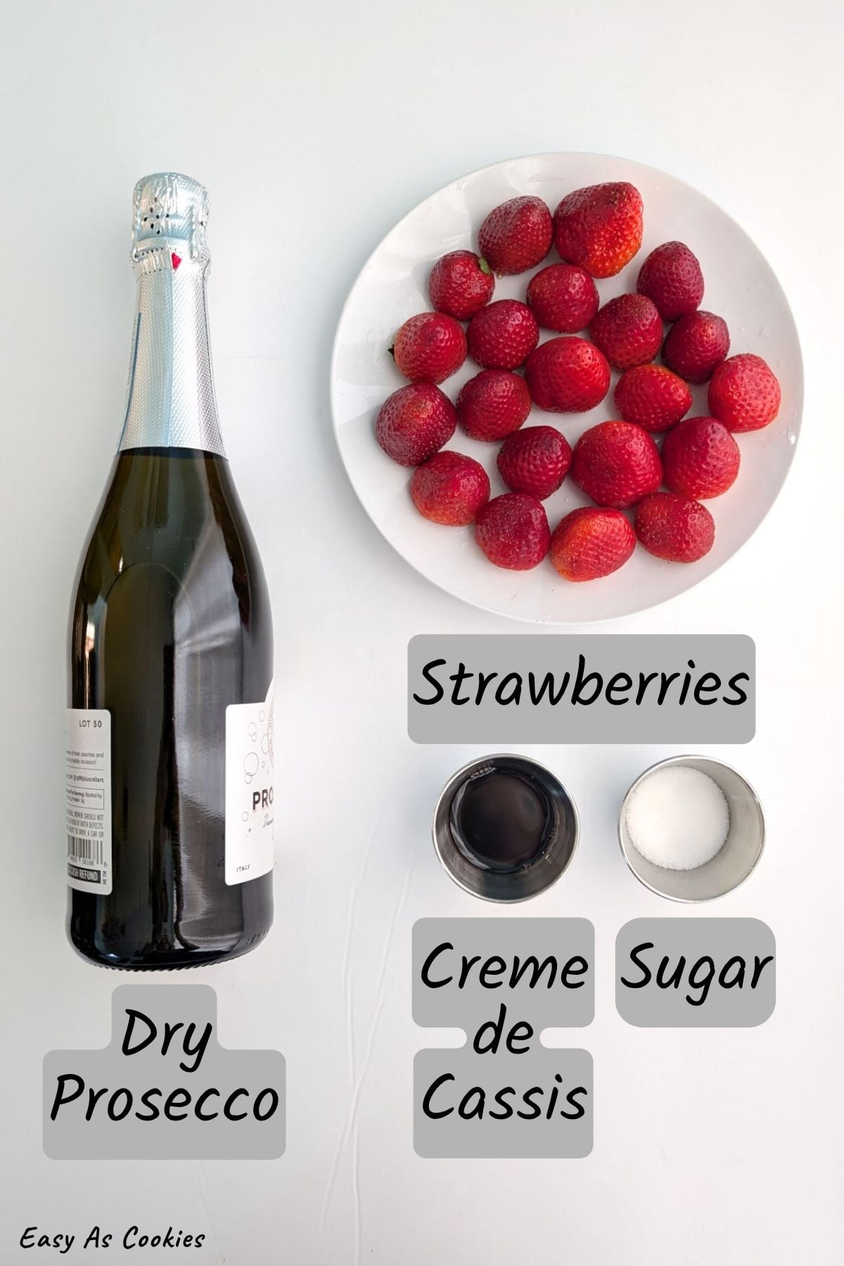 Labeled photo of ingredients for strawberry bellinis, including a bottle of dry prosecco, fresh strawberries, creme de cassis, and sugar.