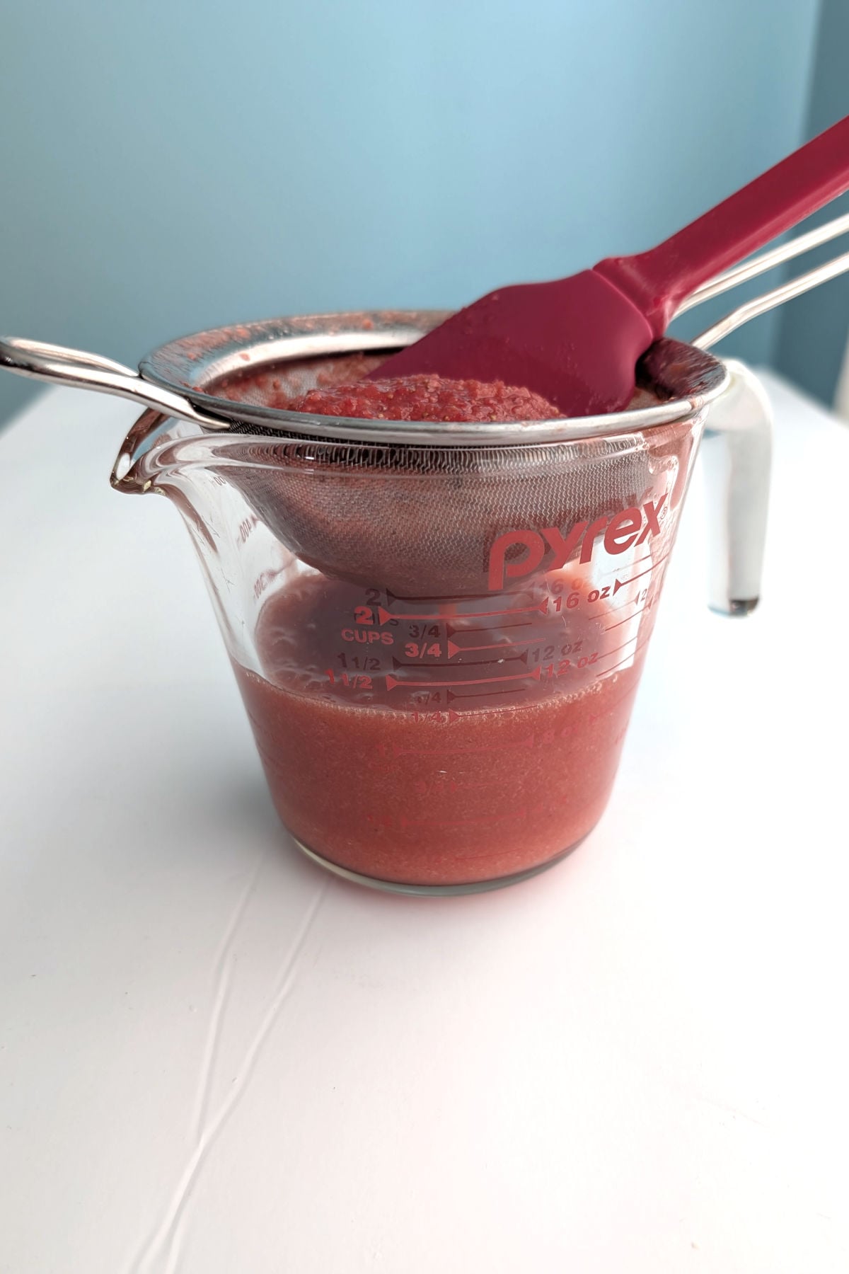 Strawberry puree in a glass measuring cup. On it rests a mesh strainer with seedy pulp, and a silicone spatula.
