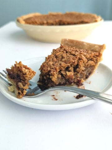 Close-up of a black bottom oatmeal pie slice showing a rich, dark filling and a textured oat crumble topping. A fork in the foreground displays a perfect bite, set against a minimalist white table and a dusty blue wall.