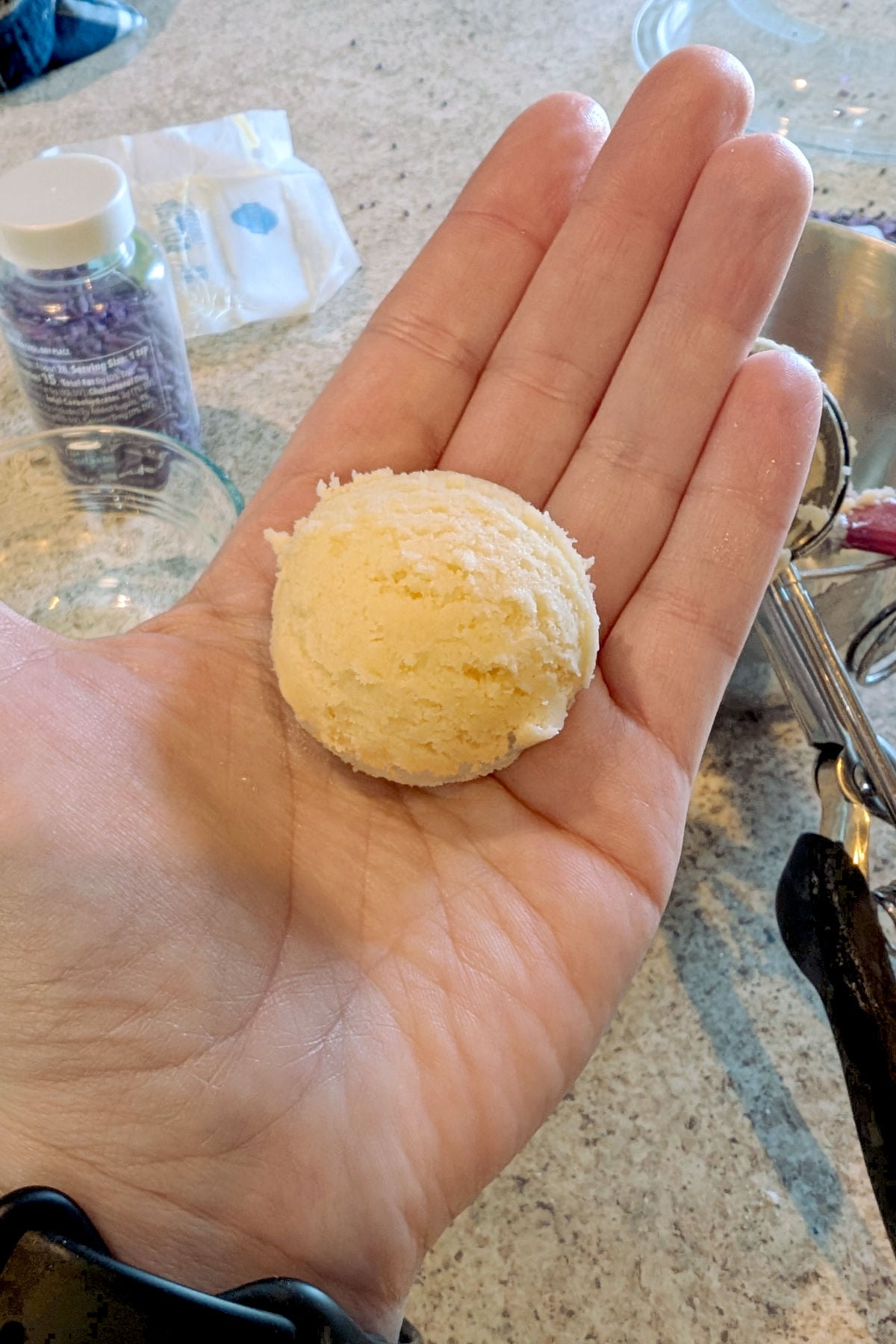 A close-up, top-down view of a hand holding a perfectly round scoop of pale, creamy cookie dough. The dough has a detailed, textured "scalloped" surface from being scooped. The background shows a granite countertop with a container of purple sprinkles, a butter wrapper, and a metal cookie scoop.