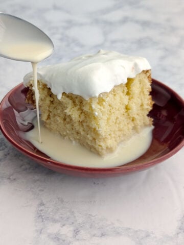 A spoon drizzles milk into a puddle next to a slice of tres leches cake.
