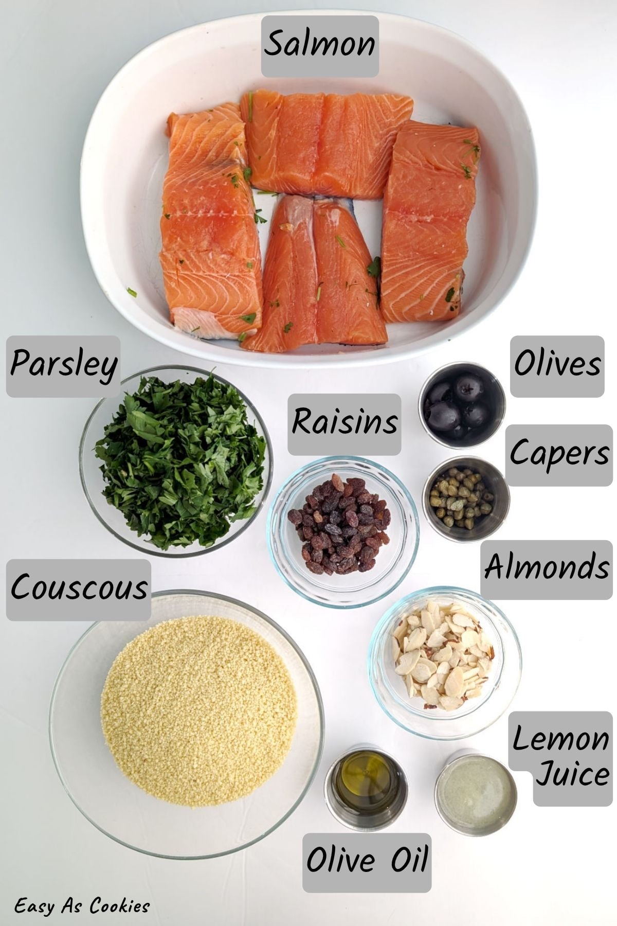 Labeled photo of ingredients for Mediterranean Salmon with Baked Couscous, including salmon fillets, parsley, couscous, raisins, olives, capers, almonds, olive oil, and lemon juice.