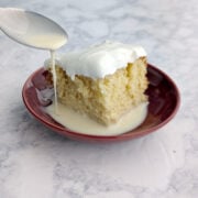 A spoon drizzles milk into a puddle next to a slice of tres leches cake.