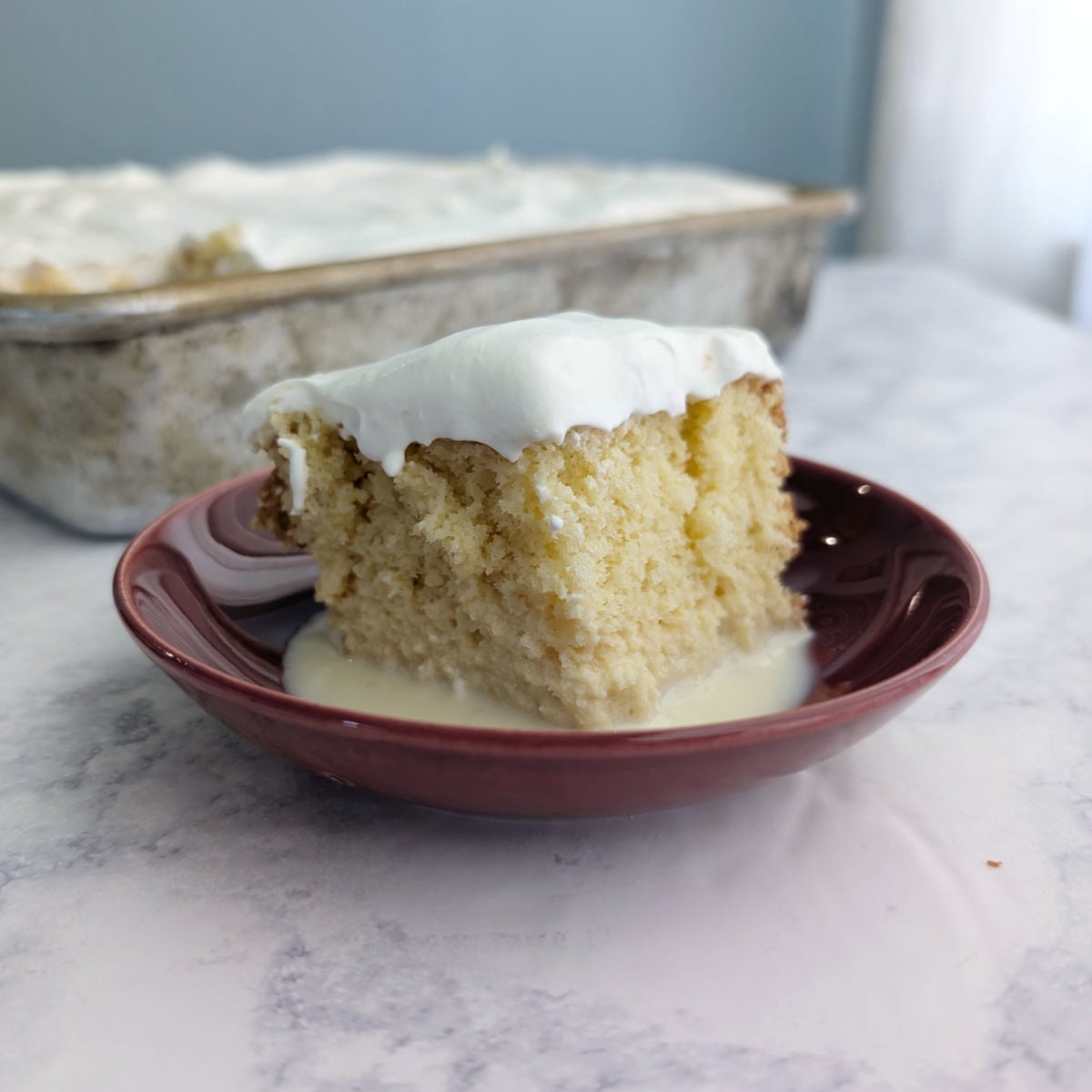 A slice of Tres Leches Cake drizzled with milk, in front of a pan.