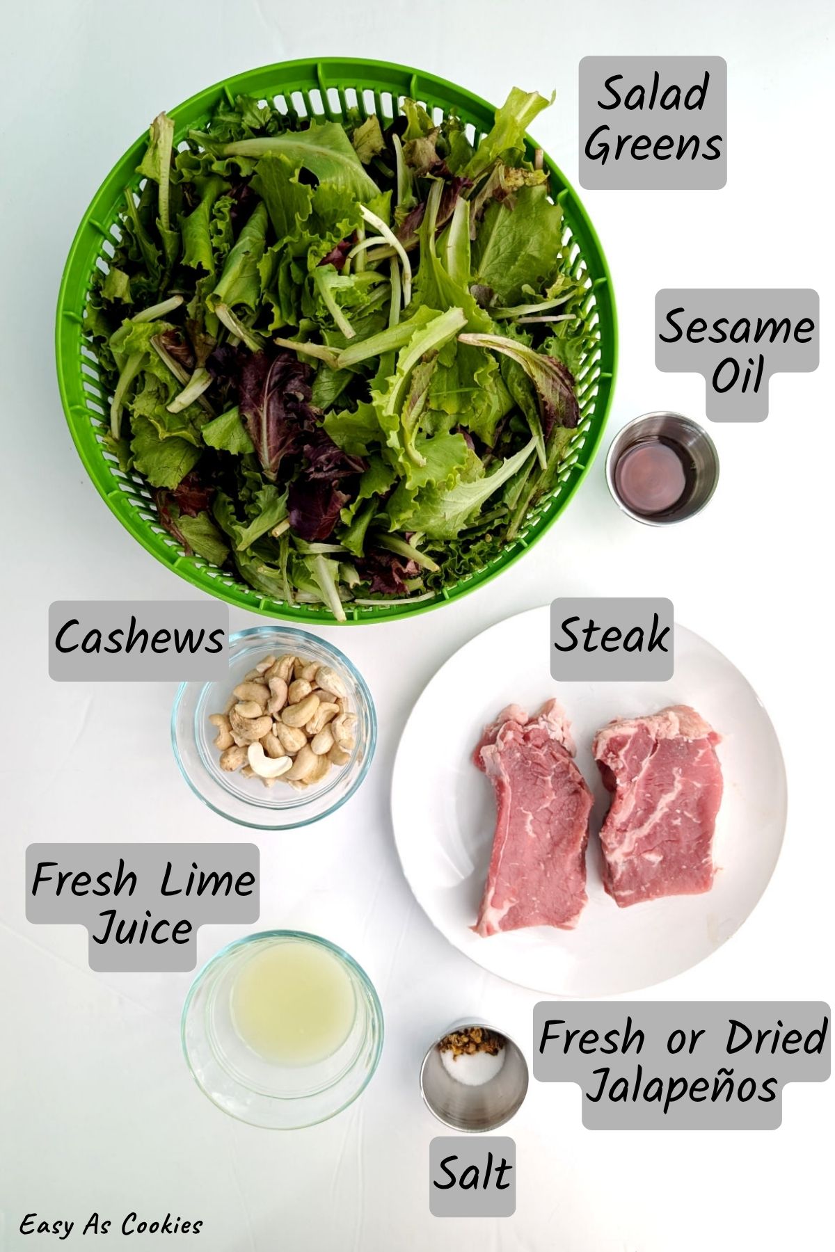 Labeled photo of ingredients for Steak Salad with Lime and Cashews, including salad greens, sesame oil, raw cashews, fresh lime juice, steak, dried jalapeños, and table salt.