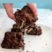 A hand holds a s'mores brownie to show the top. Other brownies are on the plate below. Text overlay reads "Easy S'mores Brownies. One Bowl. No Toasting Required. Easy As Cookies."