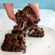 A hand holds a s'mores brownie to show the top. Other brownies are on the plate below. Text overlay reads "Gooey S'mores Brownies. Fall-Apart Soft. Easy As Cookies."
