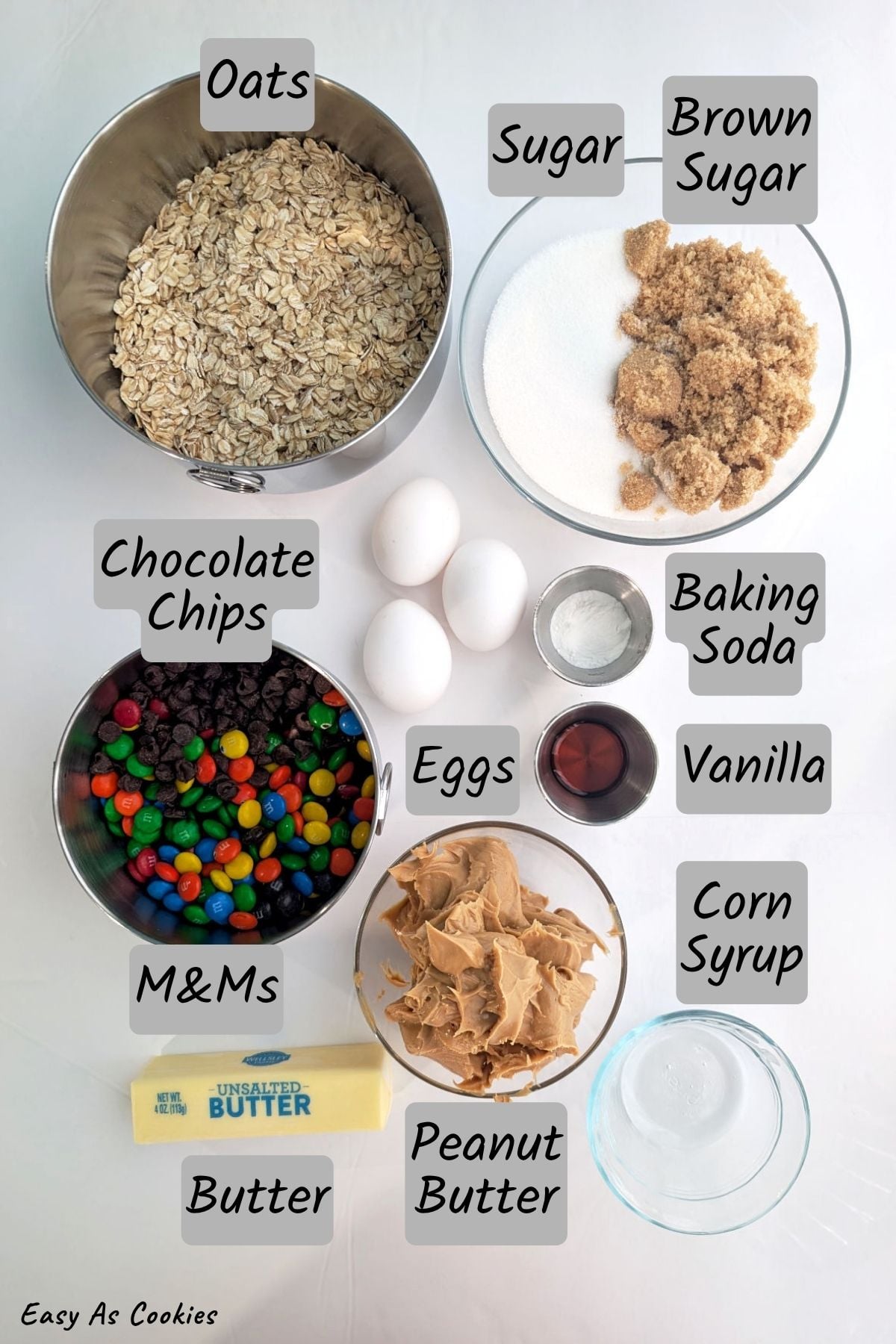 Labeled photo of ingredients for flourless monster cookies, including rolled oats, sugar, brown sugar, chocolate chips, M&Ms, butter, peanut butter, eggs, vanilla, baking soda, and corn syrup.