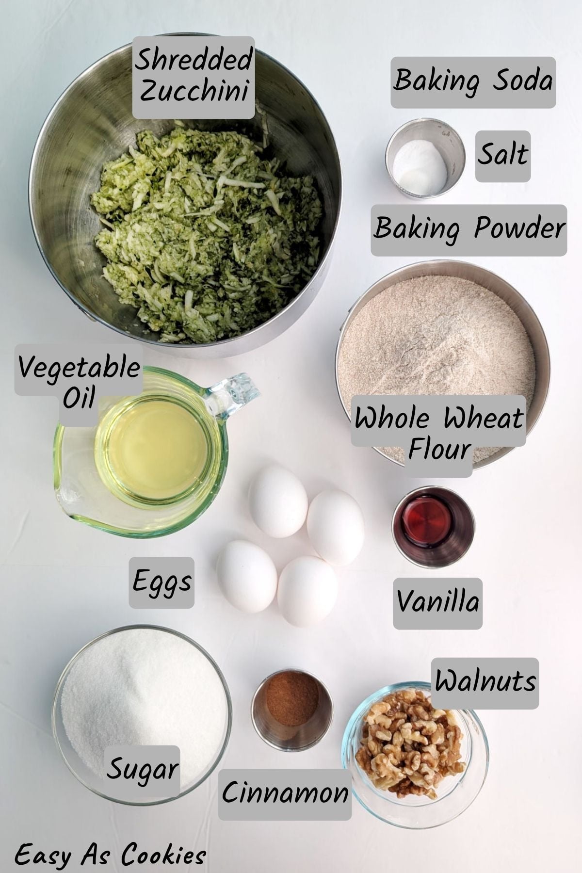 Labeled photo of ingredients for whole wheat zucchini bread, including shredded zucchini, vegetable oil, sugar, eggs, cinnamon, vanilla, whole wheat flour, baking soda, salt, baking powder, and walnuts.