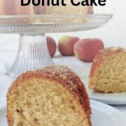 A slice of apple cider donut cake is on a plate, in front of another slice on a plate. The full cake is in the background. Text overlay reads "Apple Cider Donut Cake. Easy As Cookies."