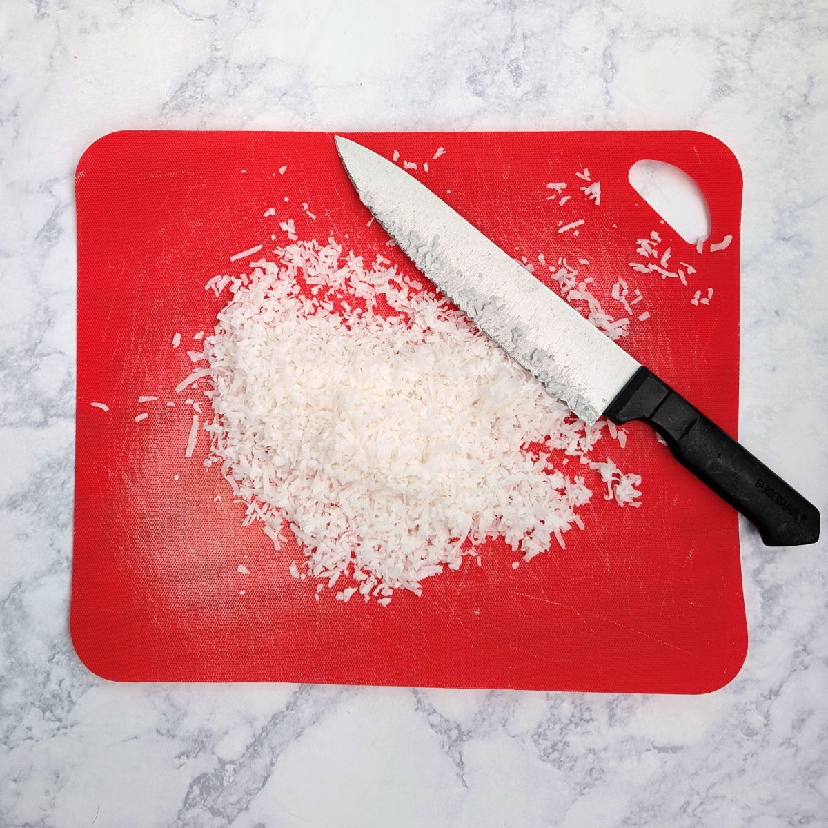 Chopped sweetened shredded coconut, on a red cutting board with a large knife next to it.