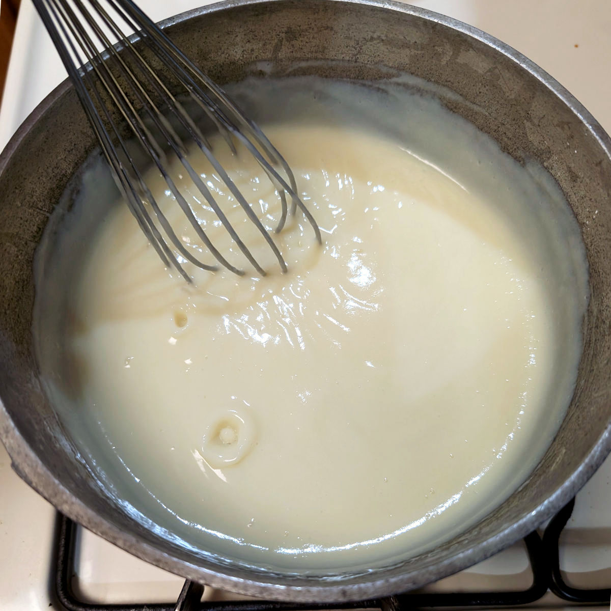 Cream pudding mixture that has thickened, in a saucepan with wire whisk. The place where a bubble popped remains visible and shows how the pudding has thickened.