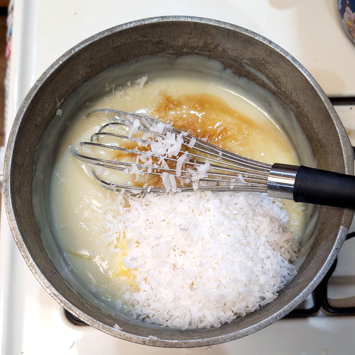 Cooked cream pie filling in a saucepan, with vanilla extract, butter, and sweetened shredded coconut on it but not yet mixed in.