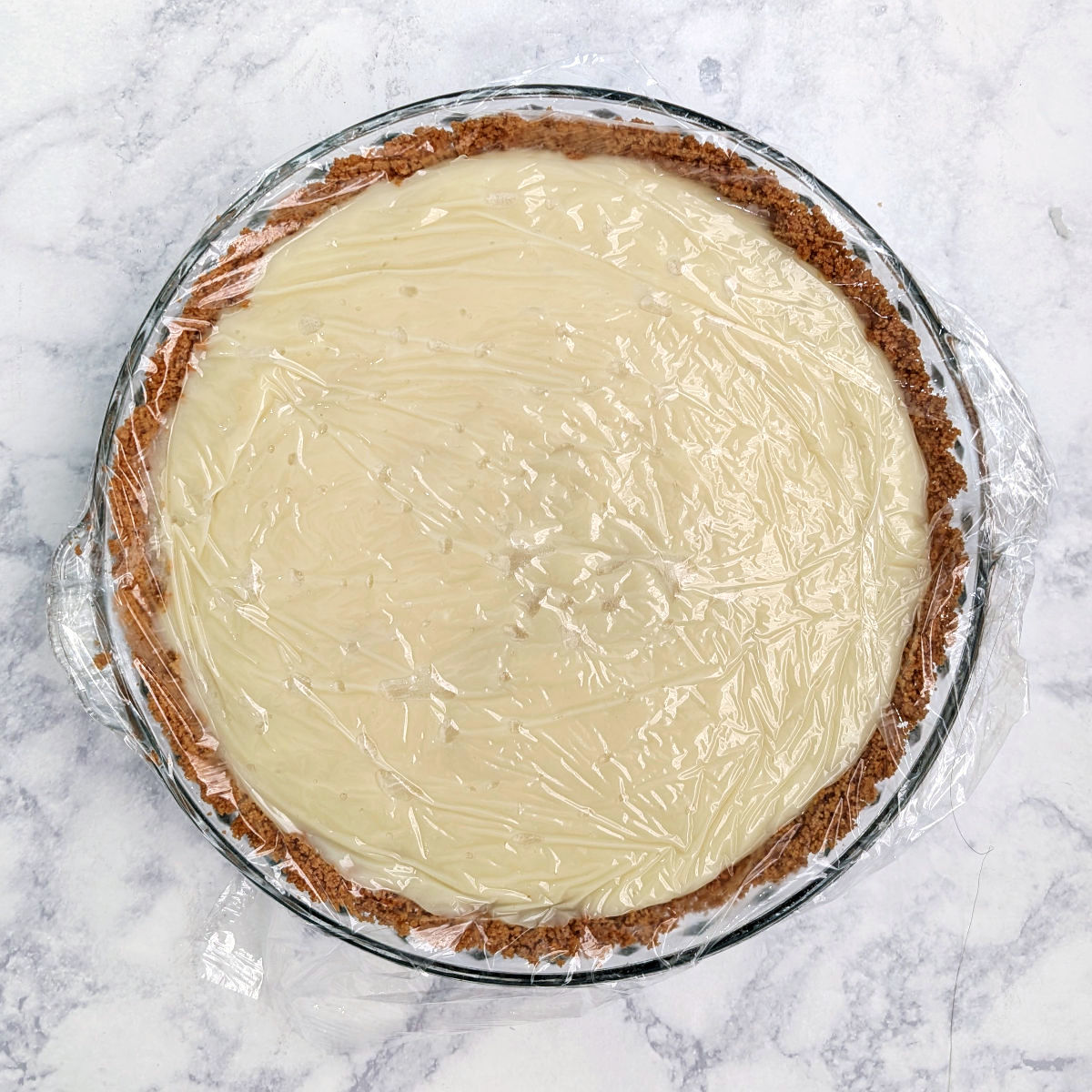 Coconut cream pie, with filling in the graham cracker crust. A layer of plastic wrap is pressed onto the surface of the pie.