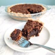 A fork with a bite of Pecan Fudge Pie rests on a plate with the slice. The remaining pie is in the background. Text overlay reads "Pecan Fudge Pie. Deep Dish Chocolate Pie. Easy As Cookies."